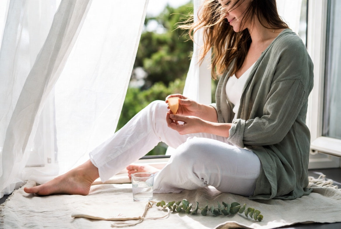 Frau sitzt am Fenster in einer gemütlichen Hausszene, hält einen Floena Menstruationstasse mit einem ruhigen Ausdruck. Natürliches Licht strömt durch die Vorhänge und schafft eine friedliche Atmosphäre, während sie den Komfort und die Einfachheit der Menstruationstasse genießt.