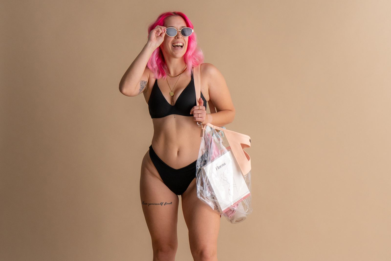 Lächelnde Frau mit pinkfarbenem Haar, die einen schwarzen Floena Leakproof Period Swim Bikini Bottom trägt und ein durchsichtiges Bag mit Floena-Verpackung hält. Sie verkörpert Komfort und Selbstvertrauen in periodentauglicher Badebekleidung und zeigt das stilvolle und auslaufsichere Design.
