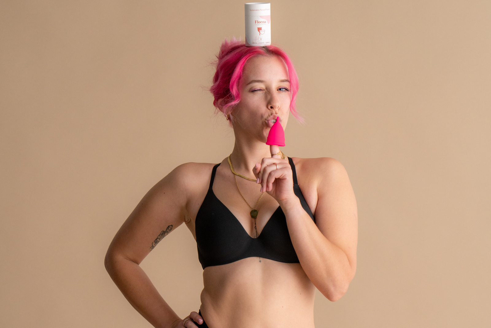 Frau mit pinkfarbenem Haar, die einen schwarzen BH trägt und spielerisch mit einer rosa Floena Menstruationstasse posiert, während sie die Floena-Verpackung auf ihrem Kopf balanciert, vor einem weichen neutralen Hintergrund und bietet diskreten, auslaufsicheren Schutz.