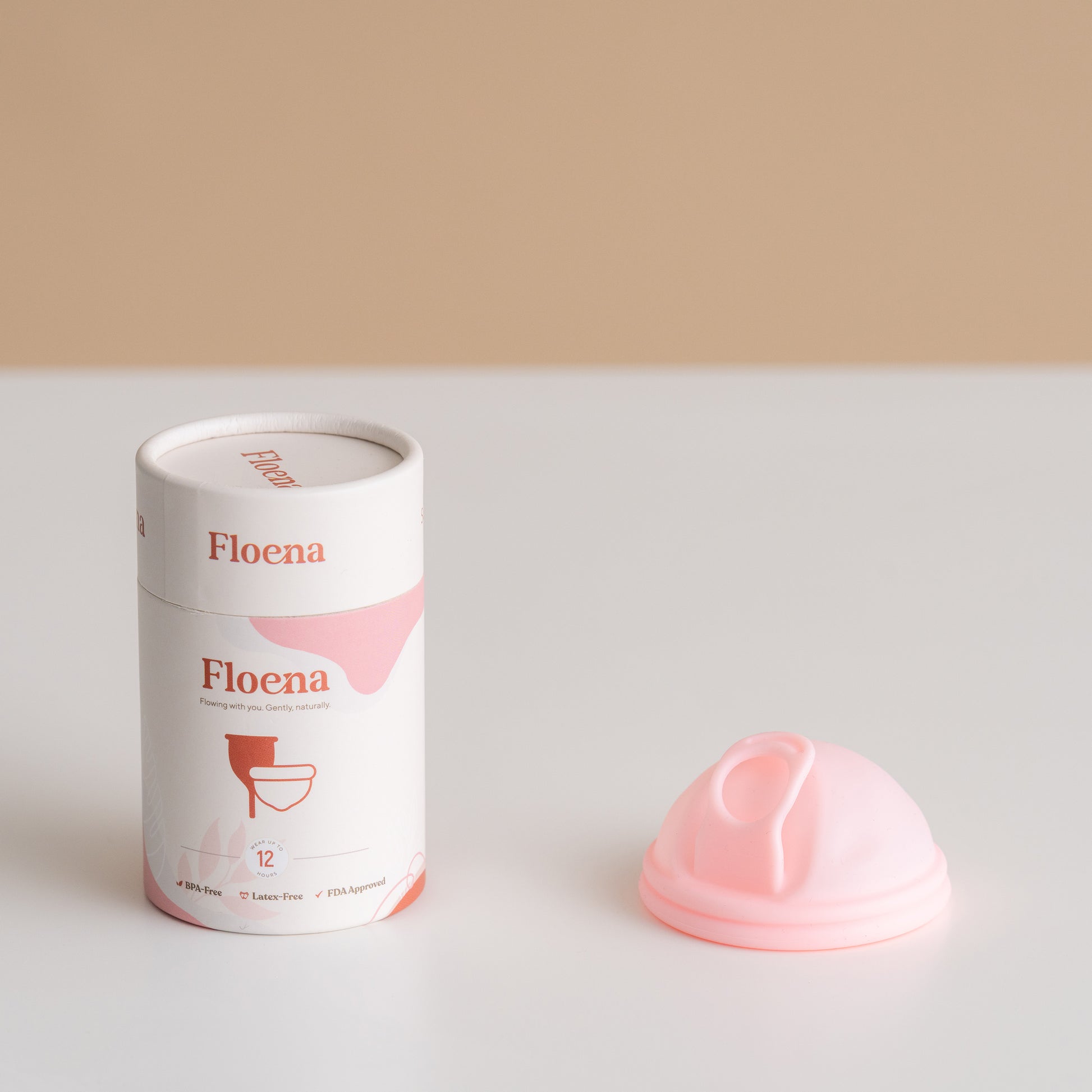 Slika prikazuje Floena menstrualni disk v minimalističnem okolju. Disk iz mehkega rožnatega silikona je postavljen poleg elegantne, moderne embalaže. Embalaža je cilindrična, z blagovno znamko v nežnem rožnatem in belem, disk pa je zasnovan za udobje in funkcionalnost med menstruacijo.
