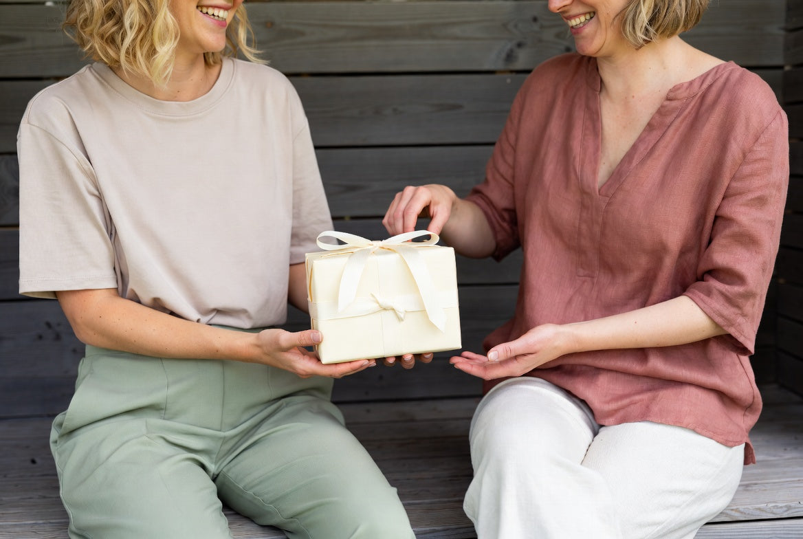 Zwei Frauen lächelnd, während sie ein eingewickeltes Geschenk austauschen, das Fürsorge, Verbindung und durchdachtes Schenken mit dem Floena Ring Cup symbolisiert, und Momente der Freude und des bedeutungsvollen Teilens hervorhebt.