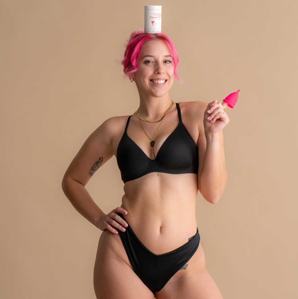 Smiling woman with pink hair wearing black underwear holds a bright pink Floena Ring Cup while a Floena product container is balanced on her head, photographed against a soft neutral background.