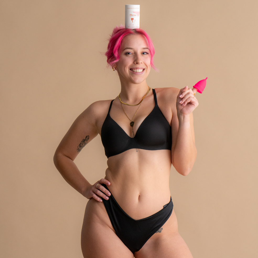 Lächelnde Frau mit pinkfarbenem Haar, die schwarze Unterwäsche trägt und eine leuchtend pinke Floena Ring Cup hält, während ein Floena Produktbehälter auf ihrem Kopf balanciert, fotografiert vor einem weichen neutralen Hintergrund.