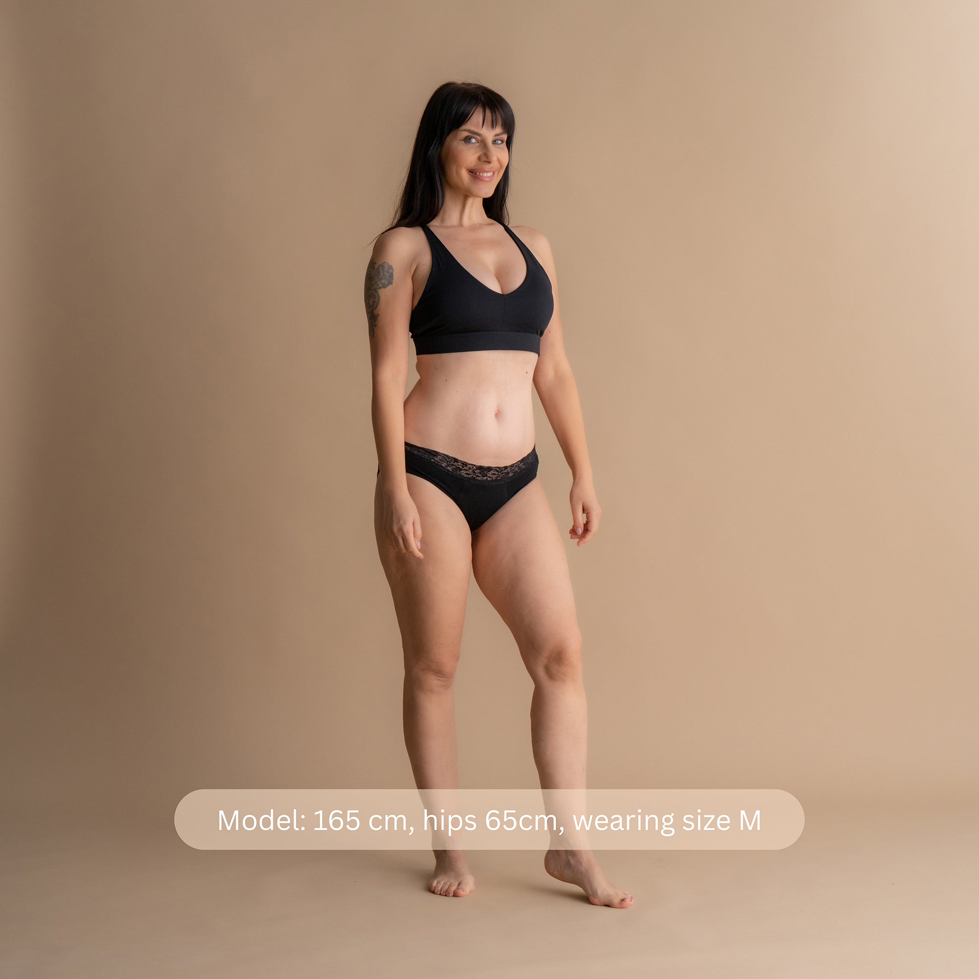 Model 165 cm tall with 65 cm hips wearing size M Leakproof Comfort Brief black lace period underwear and bra, standing against beige background with sizing info overlay