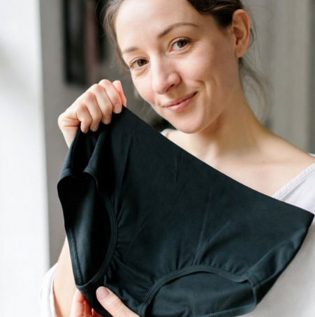 Eine Frau lächelt, während sie ein Paar schwarze Floena leakproof Periodenunterwäsche in ihren Händen hält. Der Hintergrund zeigt weiches, natürliches Licht, das durch ein Fenster strömt, und betont Komfort und Selbstvertrauen.