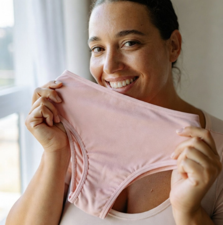 Eine Frau lächelt, während sie ein Paar weiche rosa Floena undurchlässiger Periodenunterwäsche hält und das komfortable Design zeigt. Sie steht in der Nähe eines Fensters, durch das natürliches Licht strömt.