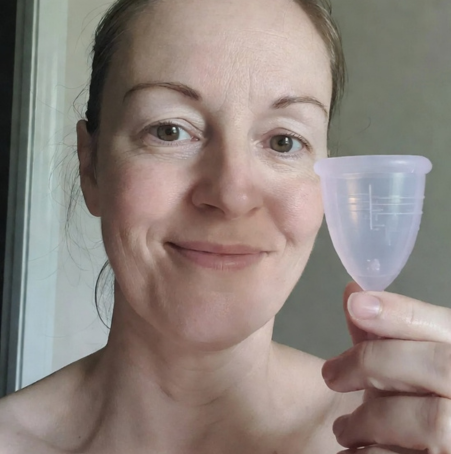 Eine Frau lächelt, während sie eine Floena Menstruationstasse in ihrer Hand hält und das praktische und umweltfreundliche Design zeigt. Sie steht in einer neutralen Umgebung mit einem selbstbewussten Ausdruck.