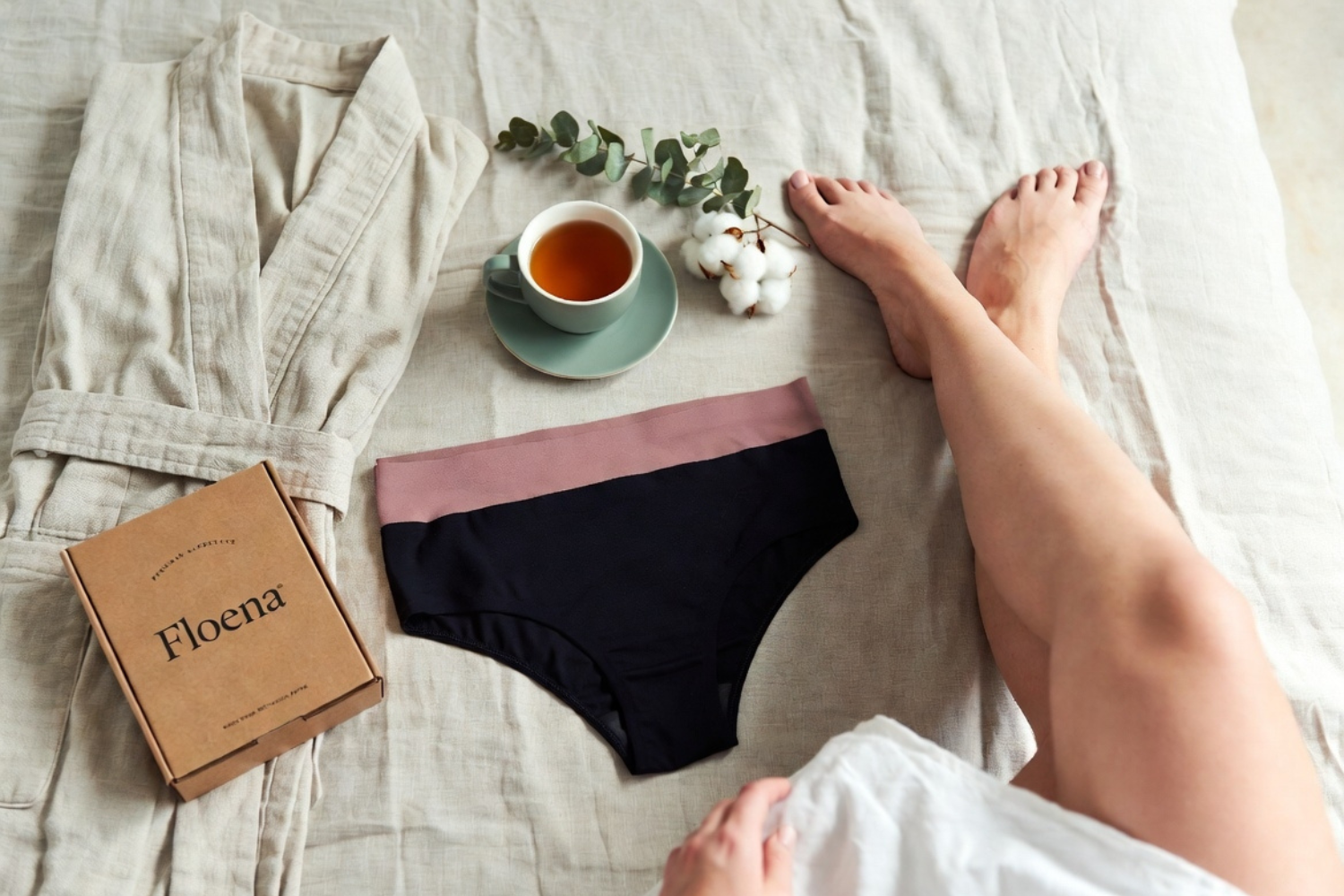 Nahaufnahme der auslaufsicheren Komfortslips in Schwarz und Pink, neben ihrer Verpackung, mit einer Tasse Tee und Baumwollblumen auf einem weichen Bett. Der Slip bietet Komfort, atmungsaktiven Stoff und auslaufsicheren Schutz.