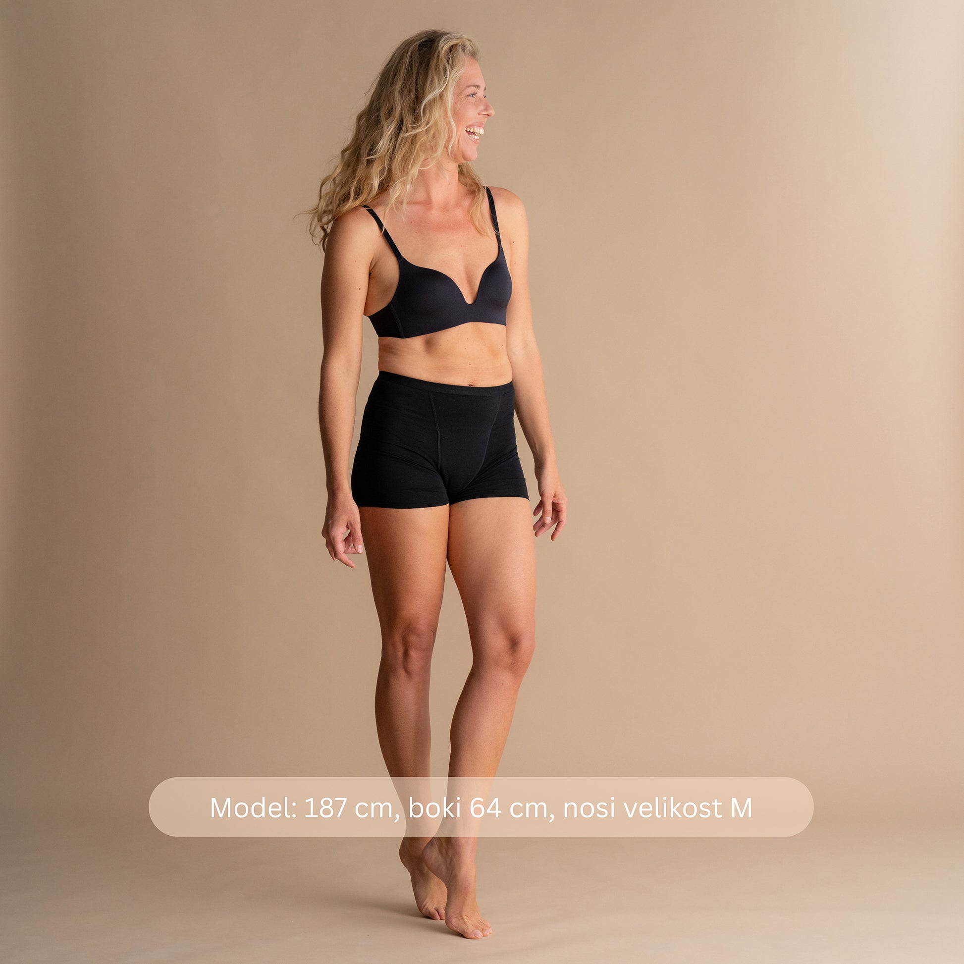 Model višine 187 cm, z boki 64 cm, nosi velikost M črnih bambusovih menstrualnih hlačk Leakproof Bamboo Sleep Short na bež ozadju