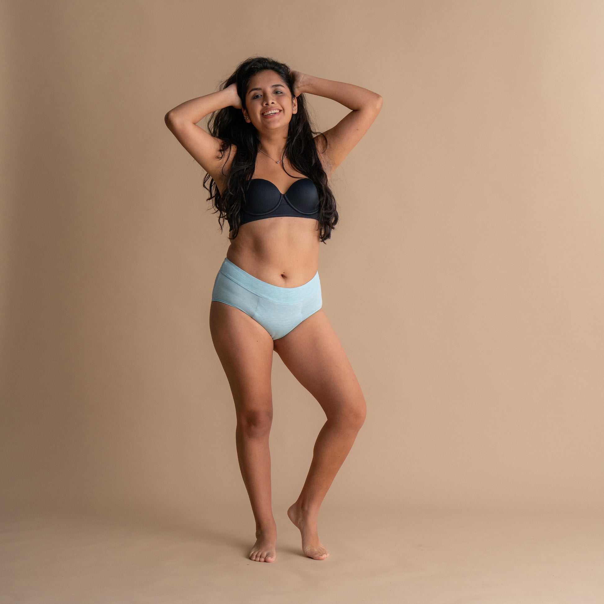 Smiling model wearing light blue Leakproof Bamboo High Waist Brief high-waisted period underwear and black bra, standing against beige background