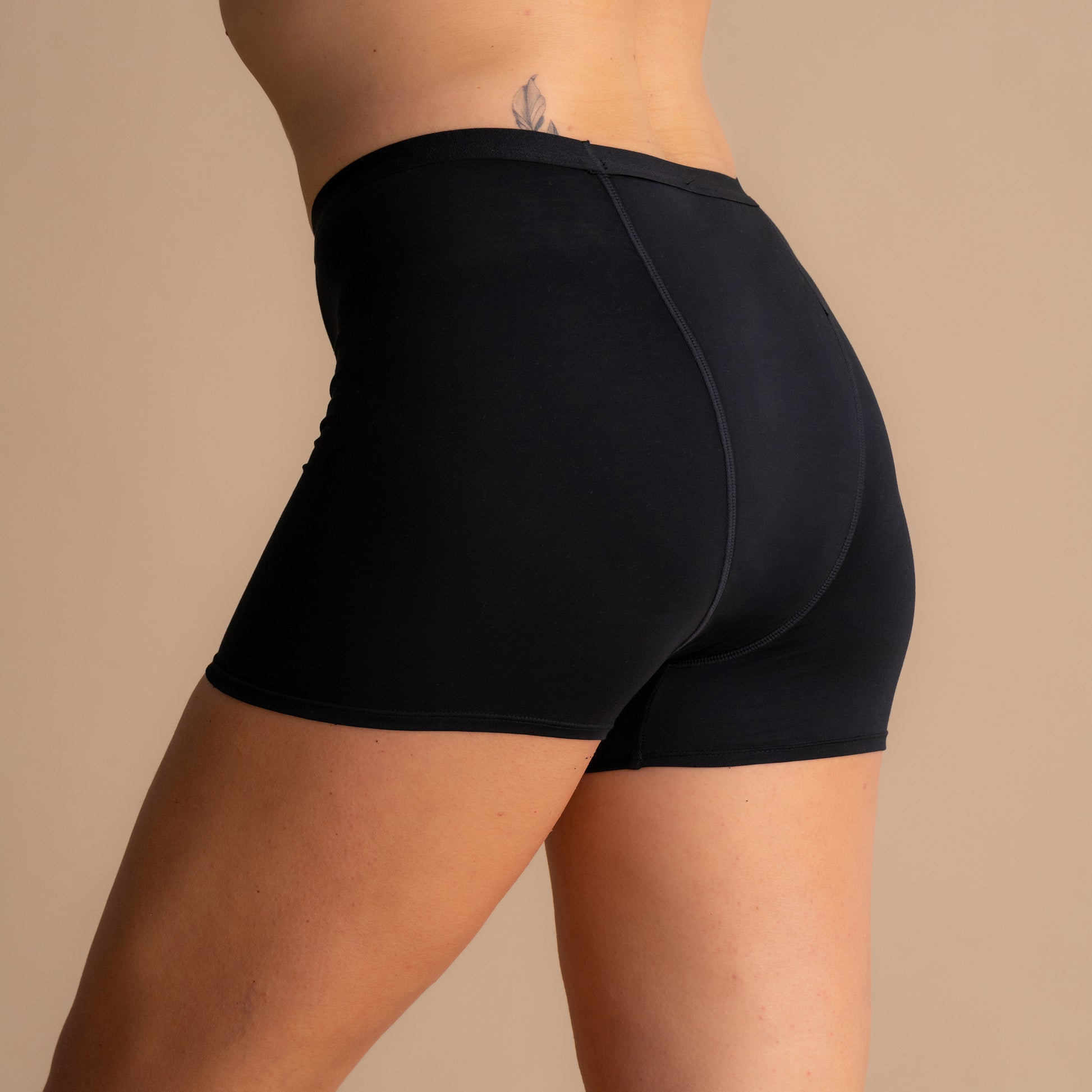 Zadnji pogled na črne Leakproof Bamboo Sleep Short menstruacijske spalne hlače na modelu, ki prikazujejo popolno prileganje, pred bež ozadjem.