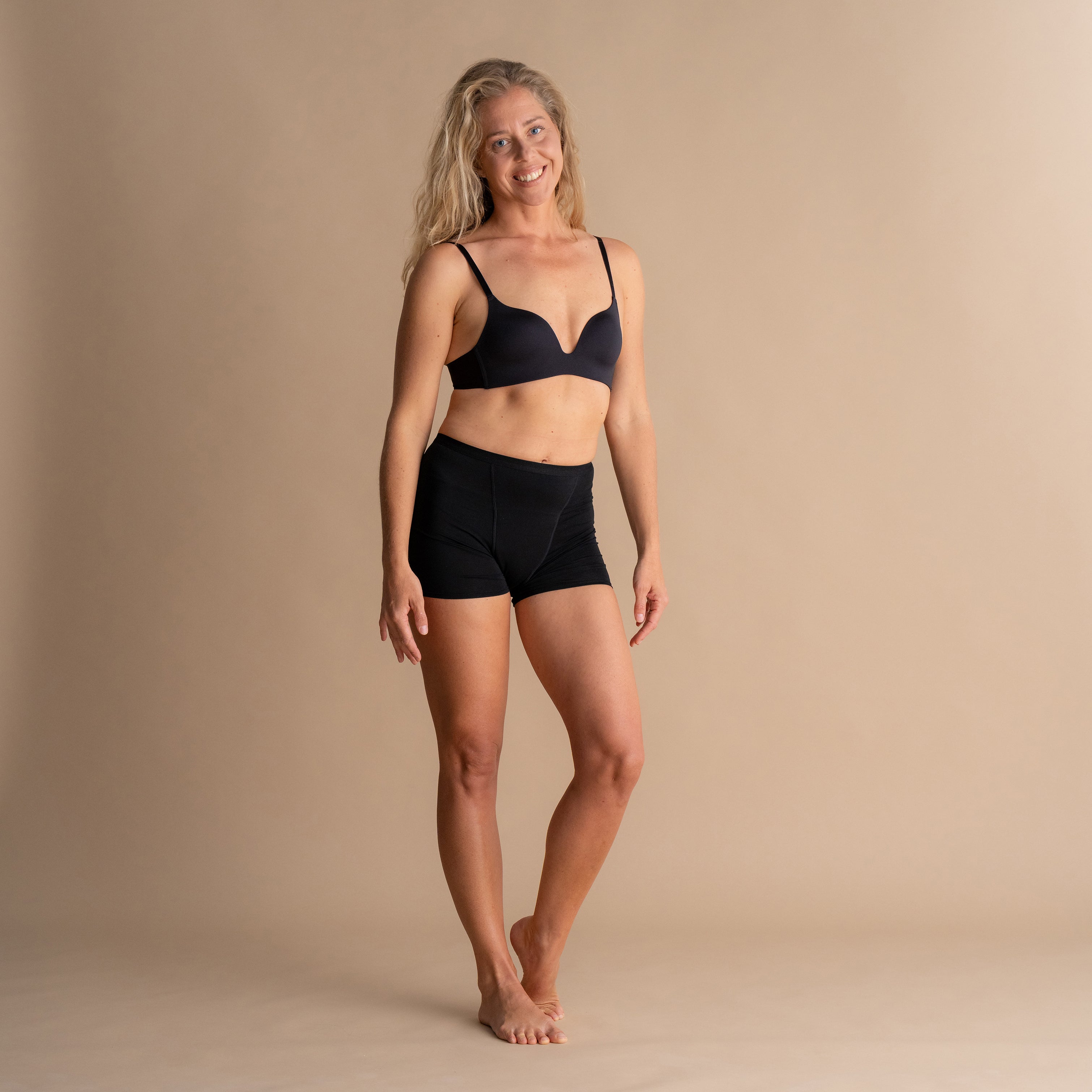 Lächelndes Model trägt schwarze Leakproof Bamboo Sleep Short Periodenschlafshorts und passenden BH, steht vor beigem Hintergrund.