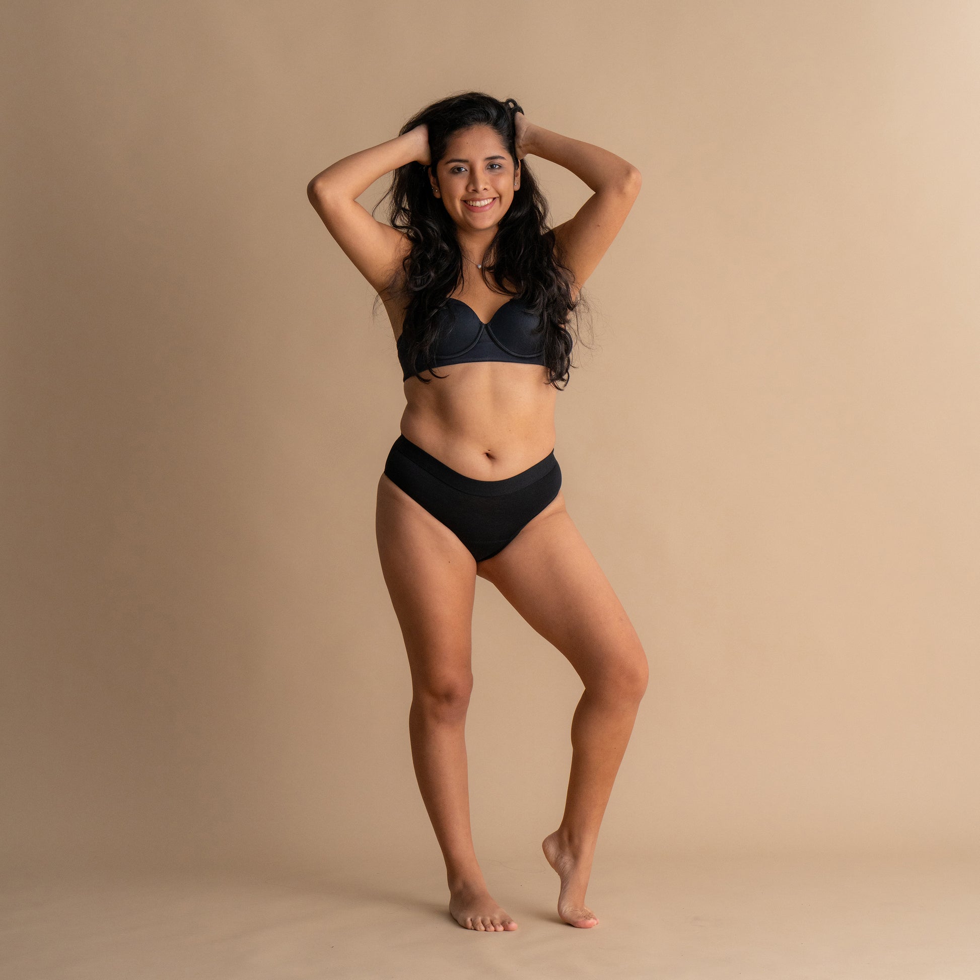 Celotni sprednji pogled modela, ki nosi Leakproof Everyday Bamboo Bikini v črni barvi. Model se smehlja in ima roke v laseh, pri čemer prikazuje udoben, mehak material in visok pas bikinija.