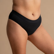 Full side view of the model wearing the Leakproof Everyday Bamboo Bikini in black, showcasing the comfortable, breathable fabric and the smooth waistband design. The model is posed naturally in a standing position, demonstrating the fit and coverage of the bikini.