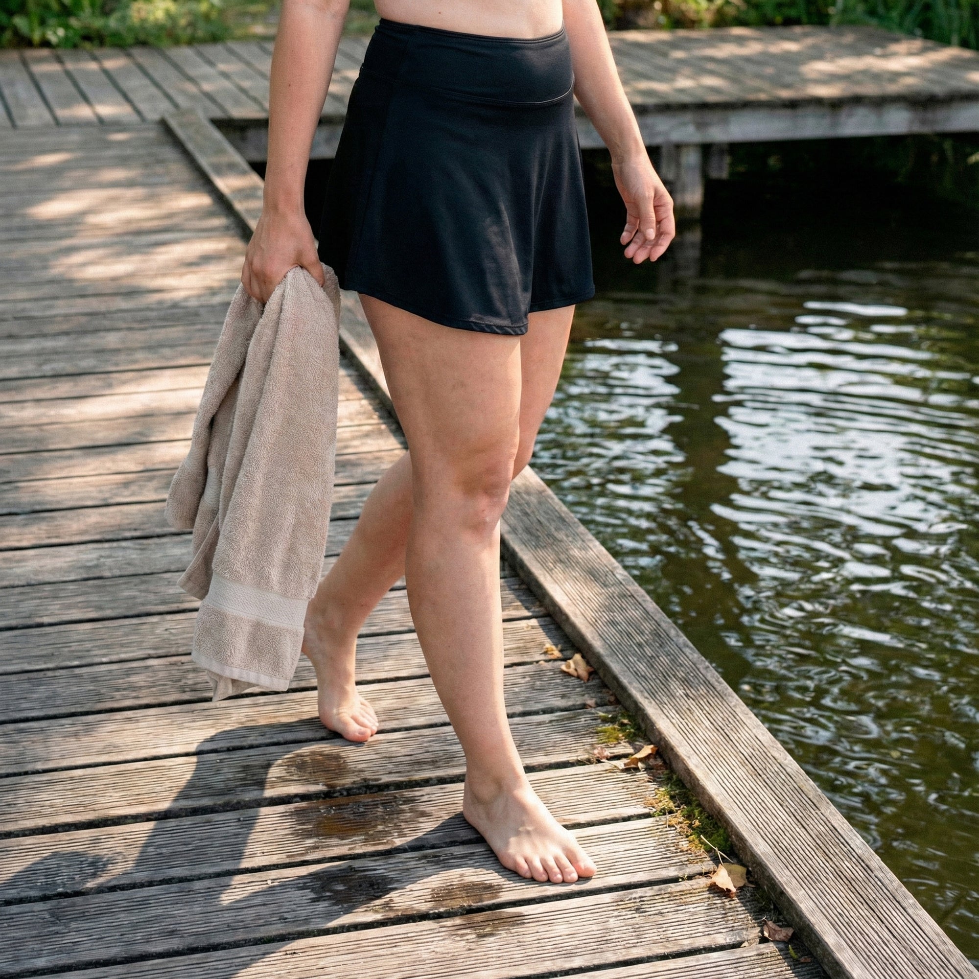 Leakproof Period Swim Skirt – A woman walks barefoot along a wooden dock by the water, wearing a Leakproof Period Swim Skirt. She holds a towel in her hand, and the serene scene highlights the comfort and style of the swim skirt in a natural setting.