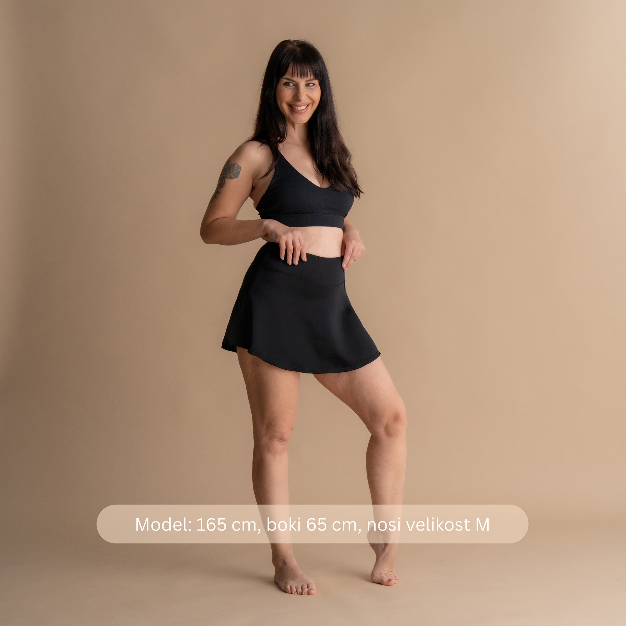 Sprednji pogled modela, ki nosi Leakproof Period Swim Skirt z ujemajočim bikini topom. Model se nasmehne samozavestno z rokama na bokih, prikazuje elegantni kroj in ženstveno silhueto plavalne krilce. Oblikovanje ponuja vgrajeno zaščito pred obdobjem, hkrati pa nudi udobje in slog za vodne dejavnosti.