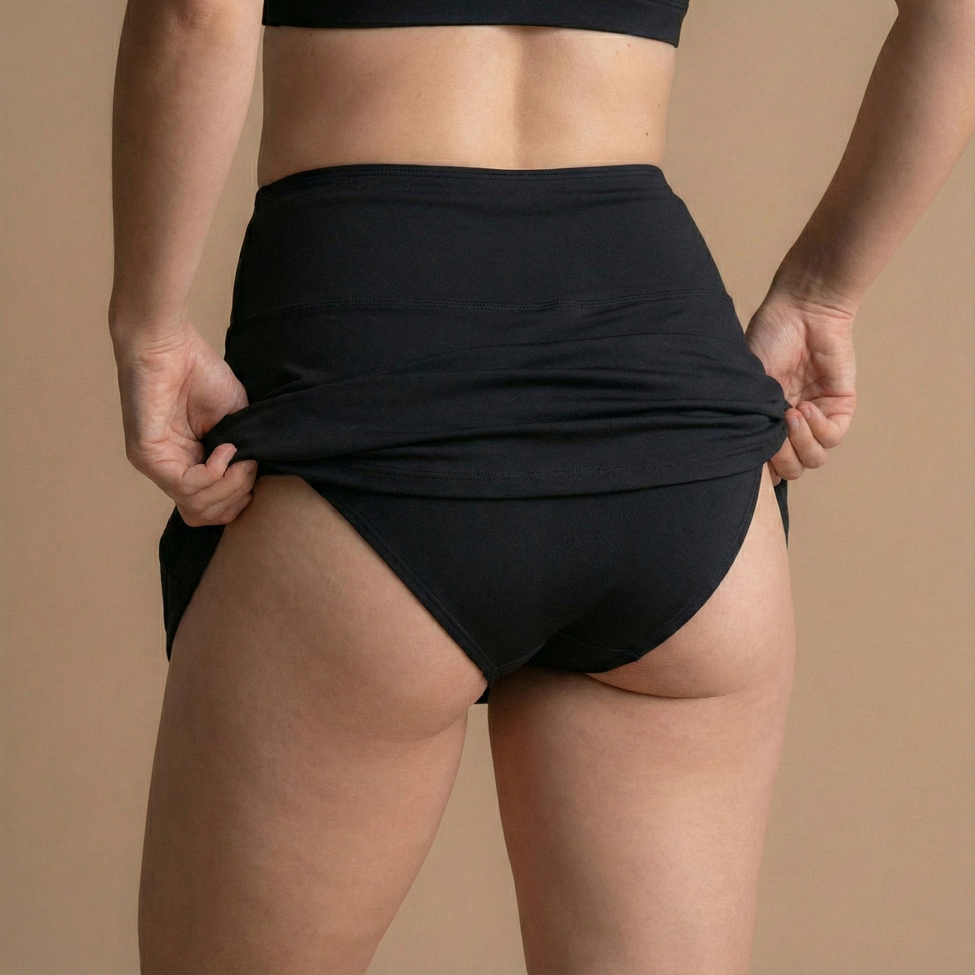 Zadnji pogled modela, ki prilagaja Leakproof Period Swim Skirt, in prikazuje diskretno, vgrajeno zaščito. Oblikovanje nudi popolno pokritost z laskavim, visokim pasom in vodoodporno tkanino, ki zaklene morebitne madeže.