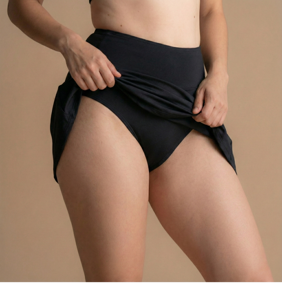 Nahaufnahme einer Person, die einen schwarzen undichten Perioden-Schwimmsrock trägt und vorsichtig die äußere Schicht anhebt, um das darunter liegende eingebaute Schutzunterwäsche zu zeigen, vor einem neutralen Hintergrund.