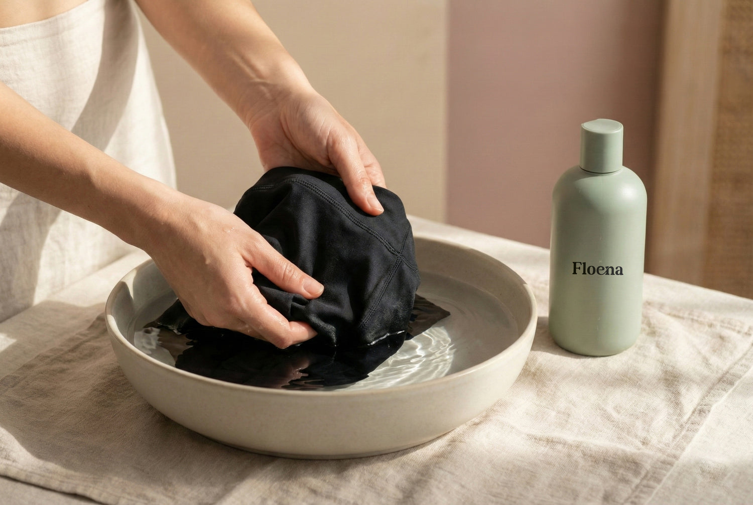Hände waschen sanft das schwarze Floena Leakproof Period Menstrual Swimwear in einer Schüssel mit Wasser, während eine Floena-Flasche daneben auf einem neutralen Stoff liegt. Das Bild hebt die Pflege und den Komfort der Floena Perioden-Badebekleidung hervor.
