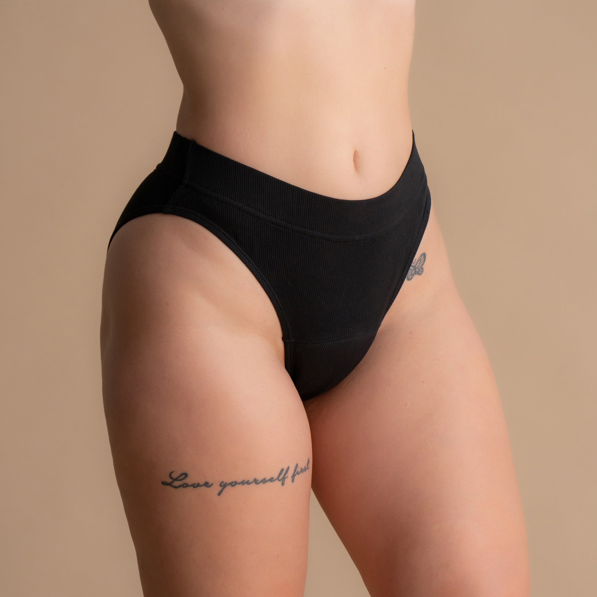 Nahaufnahme von vorne eines Modells, das schwarze leckgeschützte Rippenslips mit hohem Bund trägt – gerippte hochtaillierte Menstruationsunterwäsche vor einem beigen Hintergrund.