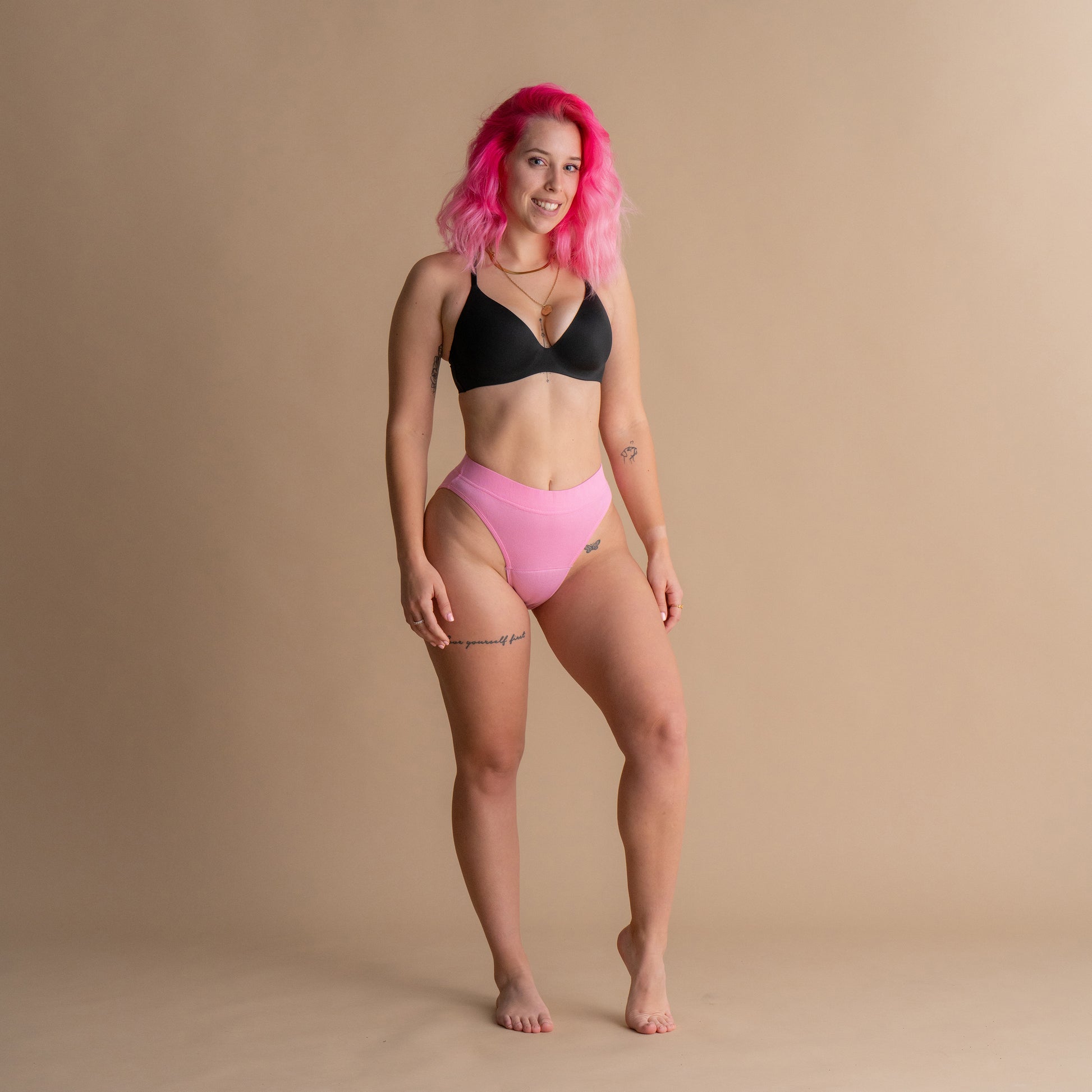 Celotni sprednji pogled modela z roza lasmi, ki nosi roza Leakproof Ribbed High Waist Brief - rebraste visoko pasne menstrualne hlačke in črn modrček na bež ozadju