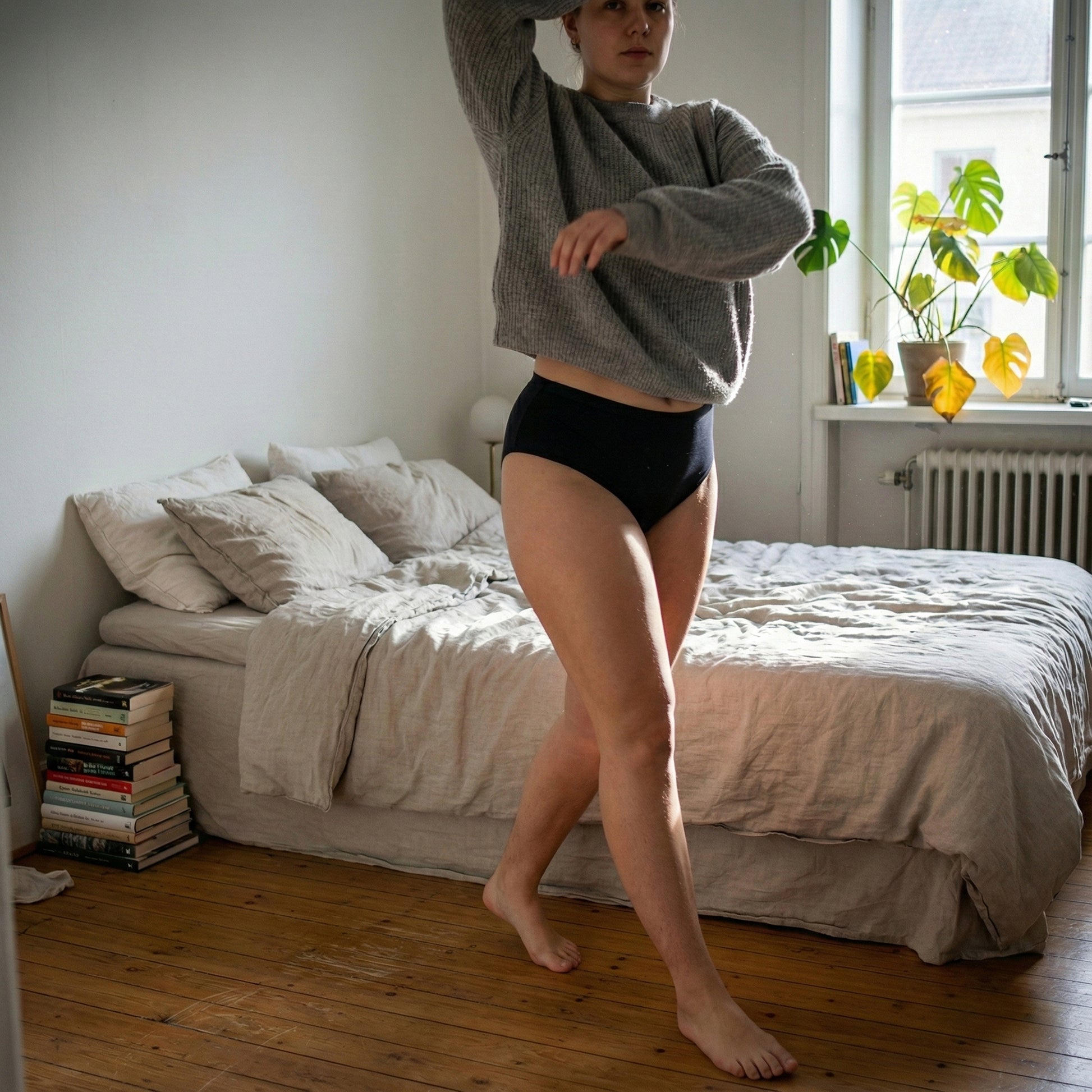 Leakproof Seamless High Waist Brief - Lifestyle slika – Slika prikazuje model v spalnici, ki nosi sivo jopo in črne brezšivne hlačke z visokim pasom. Model hodi po sobi, medtem ko naravna sončna svetloba priteka, in prikazuje udobje ter prileganje hlačk.